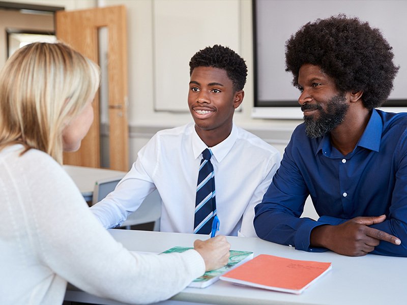 Father sat next to son, talking to a teacher in a school classroom.