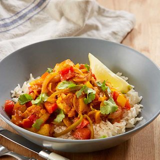 A bowl of chicken and red peppers in a spiced tomato sauce, served with a lemon wedge on a bed of rice