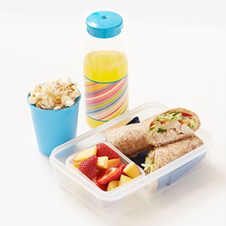 A lunchbox with a chicken wrap, chopped mango and strawberries, a pot of popcorn and a small bottle of orange juice