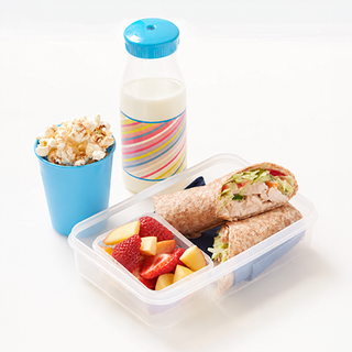 A lunchbox with a chicken wrap, chopped mango and strawberries, a pot of popcorn and a small bottle of orange juice