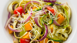 Plate of colourful pesto tagliatelle with slices of red onion, bell peppers, and squash, garnished with shredded cheese.