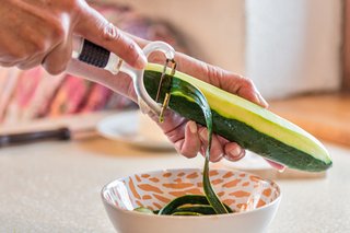 A person using a vegetable peeler to peel the skin off of a courgette and into a bowl