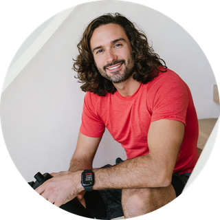 Joe Wicks is wearing a red t-shirt and sitting on a staircase.