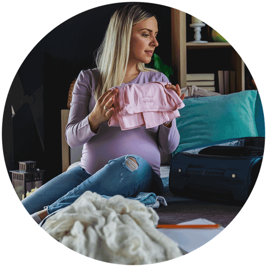 A pregnant mum to be is sitting on the bed, folding clothes ready to pack them into a small open suitcase.