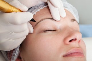 A close-up of someone wearing gloves and using a sterile needle to create permanent eyebrow definition on a young woman.