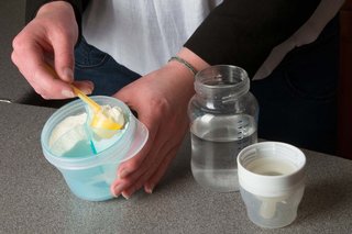 Close-up of a person holding a container of milk powder in one hand and loosely filling a scoop with powder using the other. The container is on a flat surface next to a baby bottle half-filled with water. The top of the bottle is placed upside down, with the cap covering the teat.