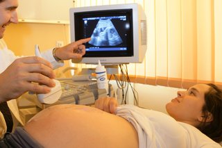 A pregnant person on a table. A radiographer is holding an ultrasound probe over their tummy and pointing to a screen.