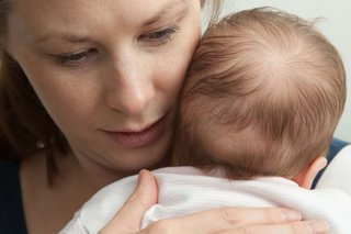 A mother holding a baby. The baby is facing the mother’s left shoulder with the side of their head resting against the mother’s cheek.