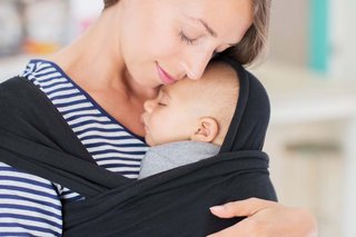 Mother holding baby against her front in a sling. The baby is held close to the mother's face with the baby's back supported.