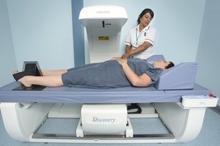 Picture of a person lying on a bed under a white scanning machine and a clinician at their side preparing them for the DEXA scan
