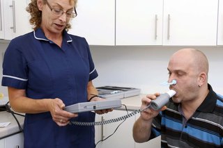 A nurse standing next to a man sitting down. The man is blowing into a tube connected to a handheld recording unit.