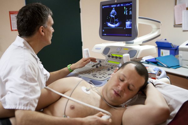 A man lying on bed with sensors and ultrasound probe on his bare chest. A technician, on left, looks at a screen