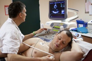 A man lying on bed with sensors and ultrasound probe on his bare chest. A technician, on left, looks at a screen