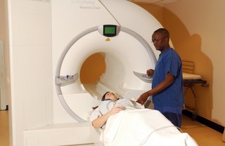 Person lying on a flat bed being moved into an MRI scanner by a radiographer operating the controls