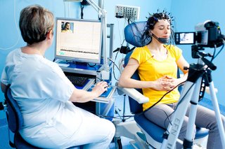 A person sitting on a chair and wearing a set of electrodes on their head while a health professional checks the EGG recording on a computer.