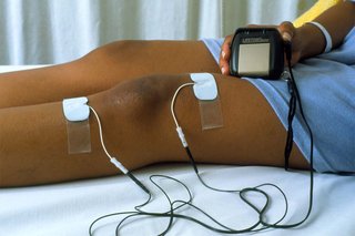 White electrode pads stuck above and below the left knee of a person lying down. The pads are attached to wires connected to a small handheld TENS machine.