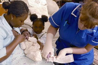 A woman holding her baby on her lap next to a nurse who is carrying out a newborn blood spot screening test.