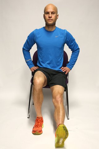 A man sitting on a chair with hands on hips, the right foot flat on the floor and the left leg straightened with the heel on the floor and toes pointing up.