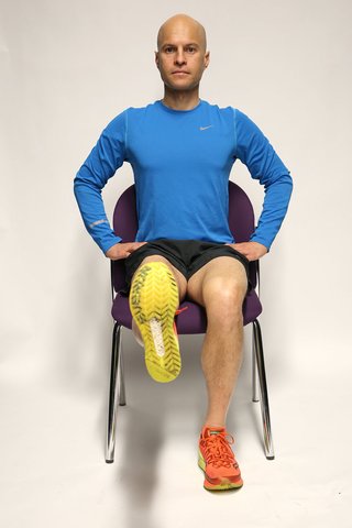 A man sitting on a chair with hands on hips, the left foot flat on the floor and the right leg straightened and raised.