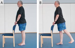 2 images showing someone doing a sequence of 2 different positions. The first image shows someone standing behind a chair with their hands on the back of the chair to support them. The second image shows someone lifting their left leg behind them, while keeping their hands on the back of the chair for support.