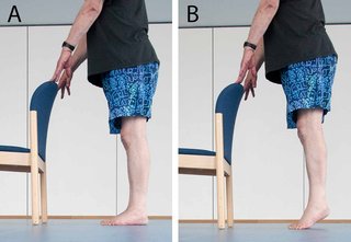 2 images showing someone doing a sequence of 2 different positions. The first image shows someone standing behind a chair with their hands on the back of the chair and their feet flat on the floor. The second image shows someone lifting their heels off the floor with their hands still supported on the back of the chair.