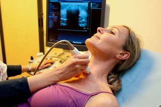 A woman lying on a bed with an ultrasound probe held against her neck.