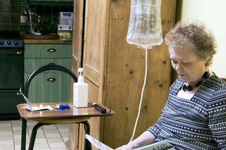 A woman having dialysis at home. She’s receiving fluid through a tube connected to a bag that is hanging above her head.