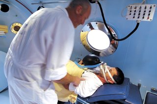 A person lying on a bed inside a hyperbaric chamber. They're wearing a mask and a clinician is attending to them.