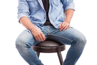 A clothed man sitting on a stool with his legs spread apart