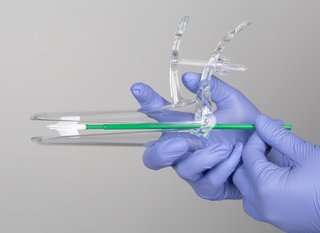 Gloved hands holding a clear plastic speculum. The speculum is a narrow tube about 15cm long. It's in 2 parts and has a hinge and handle at one end so that it can be opened slightly at the other end. Inside the speculum a plastic brush is shown with soft bristles, which can be used to collect a sample of cells when the speculum is opened.