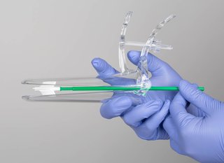 Gloved hands holding a clear plastic speculum. The speculum is a narrow tube about 15cm long. It's in 2 parts and has a hinge and handle at one end so that it can be opened slightly at the other end. Inside the speculum a plastic brush is shown with soft bristles, which can be used to collect a sample of cells when the speculum is opened.